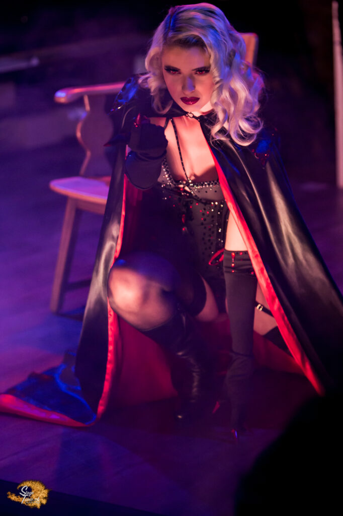 The picture shows a blonde Burlesque artist with red eyes in a vampire costume. She is alooking at an audience member with a menacing look.